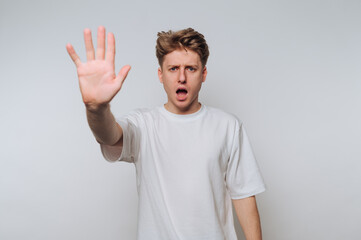 Young man with shocked expression raises hand in gesture of stopping indoors at a minimalistic space