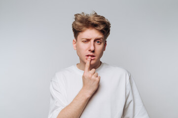 A young man with stylish hair is standing in front of a light backdrop. He is playfully winking and...