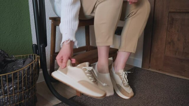 Leg view of client seated on wooden bench removing beige canvas shoes before session, with neutral pants, white sweater, green wall, coat stand, and indoor decor creating calm