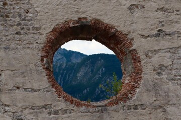 A scenic view of forested mountains through a large round hole in an old stone and brick wall. The...