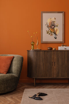Warm and eclectic interior with a retro-style olive armchair, wooden sideboard, mock up art print on orange wall. Soft lighting enhances the cozy mood.