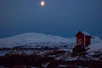 Red wooden house in traditional Norwegian style standing on the snowy beach of Telegrafbukta, southern Tromsøya, during the polar night.