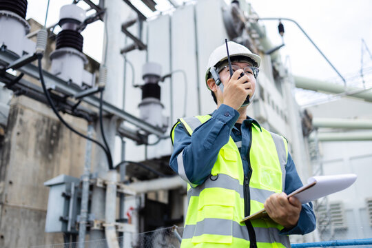Engineer is working at high voltage power substation performing inspection and check of infrastructure using radio and clipboard to ensure safety and functionality - Powered by Adobe