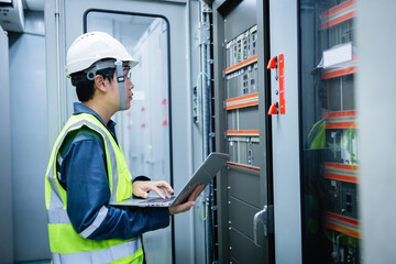 Electrical engineer in safety helmet and vest is performing inspection with laptop in substation control room near control panel focused on ensuring system functionality