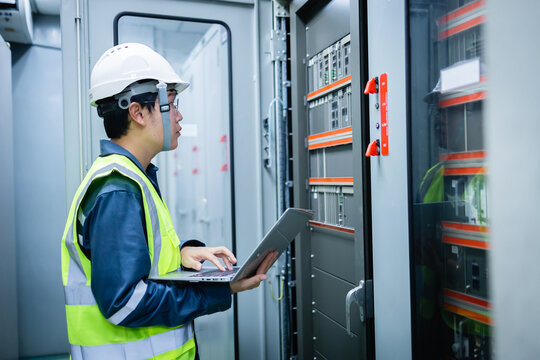 Electrical engineer in safety helmet and vest is performing inspection with laptop in substation control room near control panel focused on ensuring system functionality