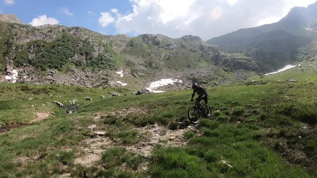 Two bikers roll through rough rocky field, patches of snow linger beside path, distant slopes fade beneath warm afternoon sun.
