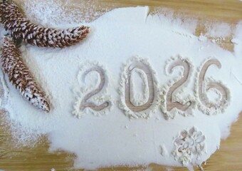 The year 2026 is written in flour on a wooden surface, decorated with two pinecones