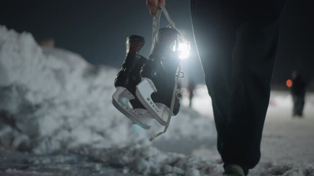 low angle close up of skater walking on snow-covered path holding black ice skate by lace, with cold night backdrop and soft lights creating serene winter atmosphere