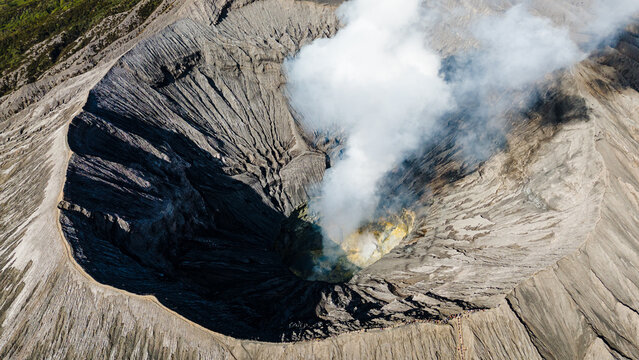 Aerial view of the stark, grey crater of Mount Bromo releasing plumes of white smoke against a backdrop of rugged terrain, Mount Bromo Volcano, East Java, Indonesia.