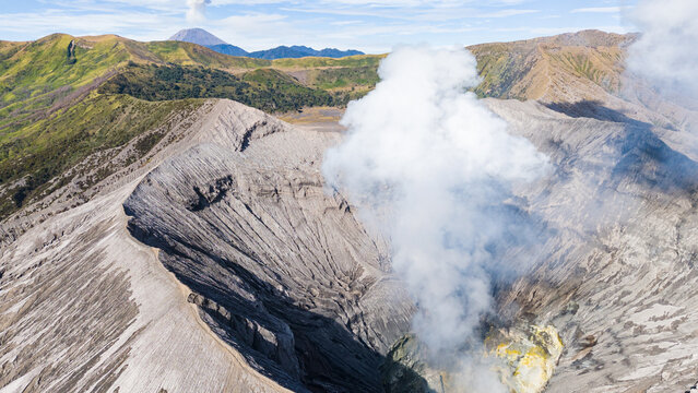 Aerial view of smoke billowing from the crater of Mount Bromo, contrasting with the arid landscape against a backdrop of distant green hills, Mount Bromo Volcano, East Java, Indonesia.