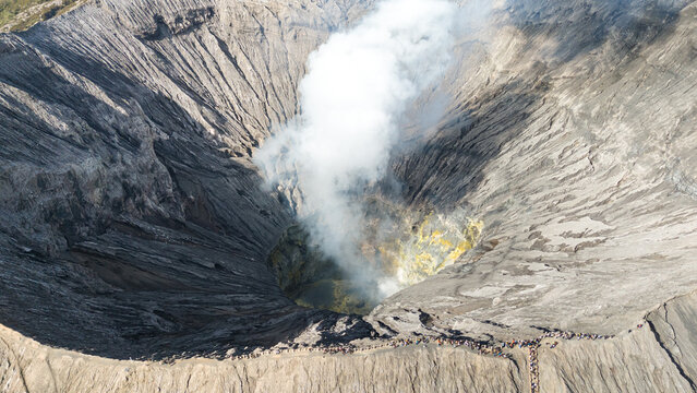 Aerial view of the Bromo volcano's crater spews forth a plume of smoke, with its steep, rocky walls forming a dramatic natural amphitheater, Mount Bromo Volcano, East Java, Indonesia.