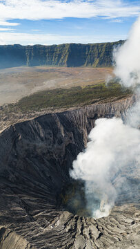 Aerial view of the rugged terrain and smoking crater of Mount Bromo, a symphony of greys and browns, veiled in ethereal white plumes, Mount Bromo Volcano, East Java, Indonesia.