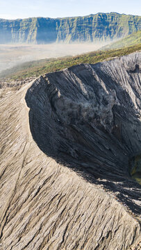 Aerial view of stark, textured volcanic slopes descend into a vast caldera, with rugged cliffs in the distance, Mount Bromo Volcano, East Java, Indonesia.