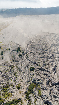 Aerial view of a rugged landscape, etched with winding paths and sparse vegetation, unfolding beneath a hazy sky near Mount Bromo Volcano, East Java, Indonesia.