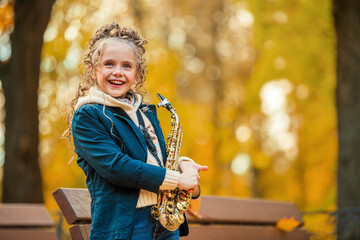 Young girl standing with saxophone in autumn park. Happy child learning music and art in nature.