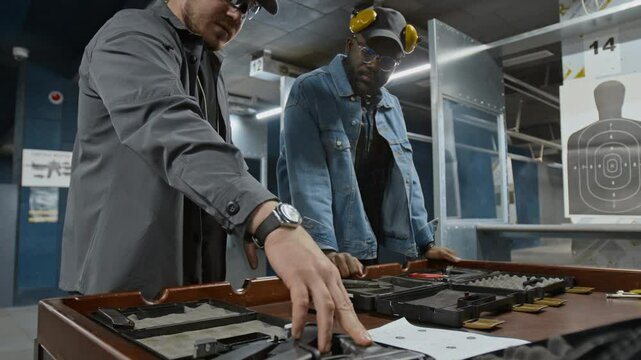 Medium closeup of young Caucasian male shooting coach instructing African American customer before practice, demonstrating assortment of handguns in padded storage cases, explaining properties