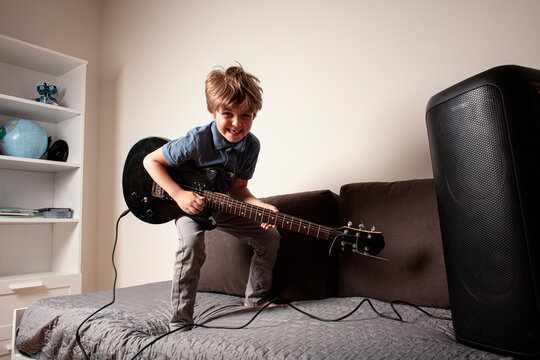 Happy boy playing electric guitar in his bedroom, sitting on the bed, laughing and enjoying music through a big speaker. Fun and creativity. Little rock star playing guitar. - Powered by Adobe