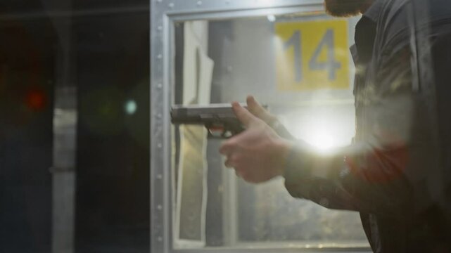 Backlit tilting closeup of hands and face of amateur civilian marksman in baseball hat and earmuffs loading bullets into magazine, inserting in handgun, aiming at target and shooting