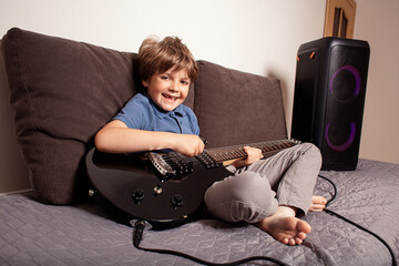 Happy boy playing electric guitar in his bedroom, sitting on the bed, laughing and enjoying music through a big speaker. Fun and creativity. Little rock star playing guitar.