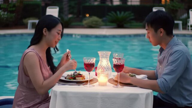 Asian couple having dinner with wine by the pool at evening
