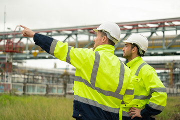 Engineers wearing safety gear discussing work at an oil refinery plant. Industrial background with metal structures and pipelines. Professional teamwork and communication in the energy indust
