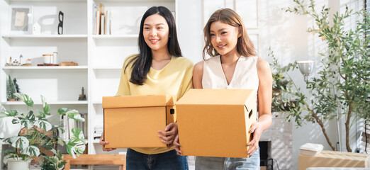 Happy Asian girl friends holding cardboard boxes moving to the new home. LGBT couple smiling,...