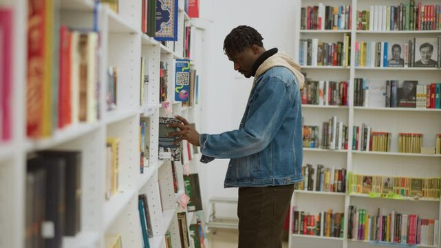 Man in denim jacket browsing bookshelves in bookstore, reaching for book while focused on selection, capturing the joy and concentration of choosing reading material in vibrant environment