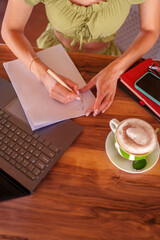 Woman drawing on notepad with pencil next to laptop and cappuccino on wooden table, working remotely from cafe