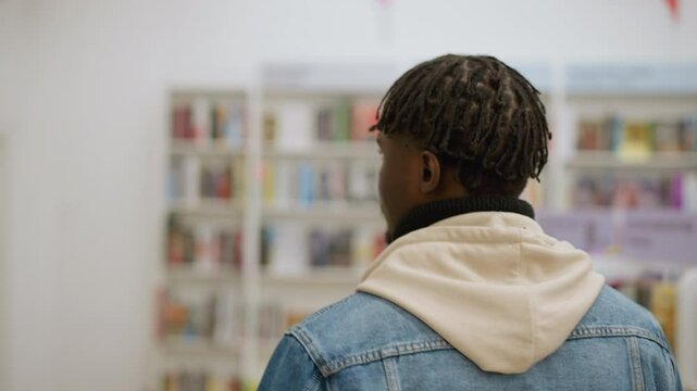 Back shot of man with dreadlocks dressed in denim jacket and hoodie, browsing books in store, with colorful bookshelves and bright lighting in background, calm and relaxed atmosphere