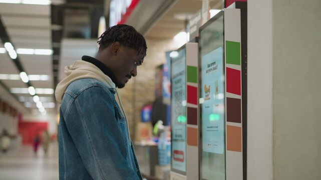 Close-up shot of young person in denim jacket and hoodie, interacting with self-service kiosk in food court. Brightly lit, digital menu displayed with food options, background showing a lively mall