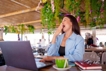 Tired woman yawning at a cafe, working on a laptop with a coffee and books nearby, surrounded by...