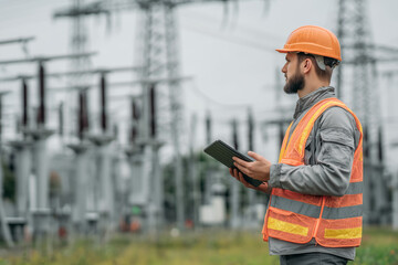 Obraz premium An electrical engineer inspecting a power substation in the field, wearing a hard hat and reflective vest, holding a tablet, high-voltage towers in the background