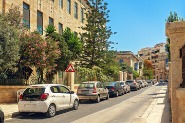 Quiet European street with parked cars along a metal fence, tropical plants, old buildings, and a...