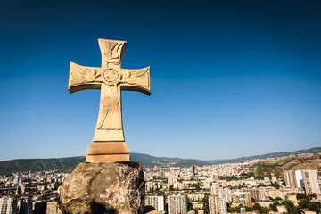 Stone cross monument overlooking Tbilisi city under bright sunlight, symbolizing faith and culture...