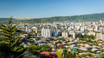 Fototapeta premium Panoramic view of Tbilisi cityscape with green hills and clear blue sky, capturing urban life against natural mountain backdrop in Georgia