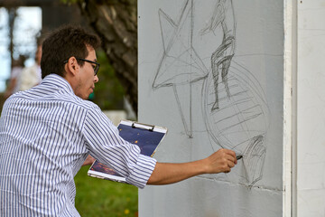 Young asian male artist sketching with pencil on outdoor wall. Focused expression, striped shirt,...