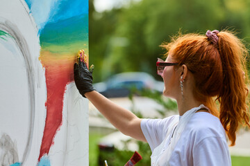 Young woman with red hair paints large canvas outdoors. Bright colors dominate, with focus on...