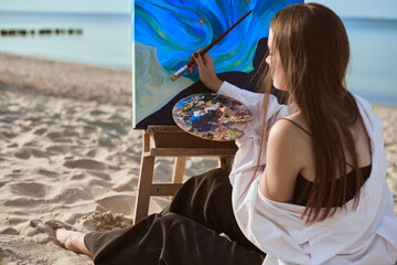 Young woman artist paints by sea, blending colors on palette. Her serene expression reflects...