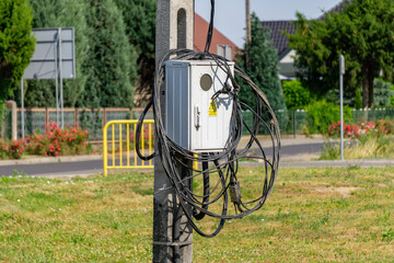 Electrical box on a pole with hanging, unsecured cables; residential area and greenery in the...