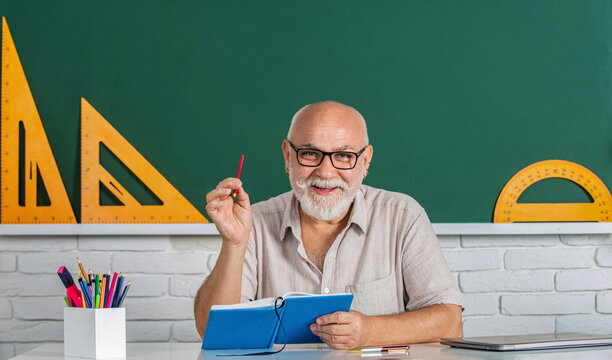 Portrait smiling man in glasses sit at desk. Happy school male teacher book. Portrait male teacher in classroom reads book. Old man teachers on green board. School male teacher reads book