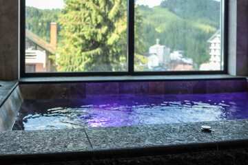 Indoor spa pool with glowing purple lights and stone edge, large window with mountain and forest view. Modern and peaceful wellness space
