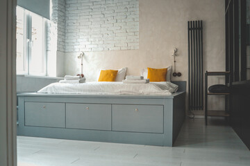 Modern minimalist bedroom with white brick walls, a large grey bed with storage drawers, yellow accent pillows, white linens, and soft daylight from a large window