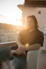 Young man holding closed book resting hands rattan chair balcony serious gaze warm sunlight glow...