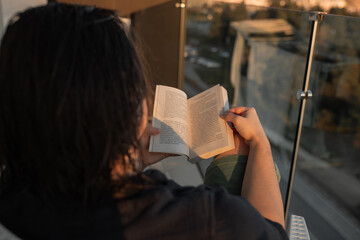 Wet-haired man seated white woven chair reading paperback book from over shoulder perspective....