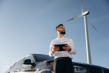 Standing by the car, holding digital tablet. Engineer in field checking on wind turbine production