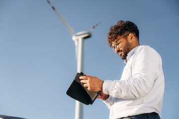 Side view, holding digital tablet. Male engineer in field checking on wind turbine production