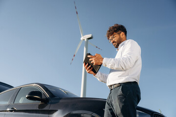 Side view, holding digital tablet. Male engineer in field checking on wind turbine production