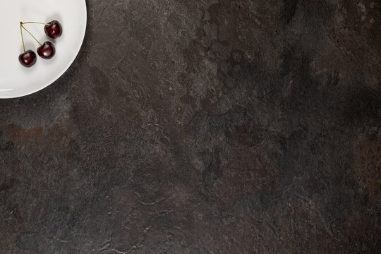 Fresh Dark Sweet Cherries on White Porcelain Plate with Slate Background. The rest of the frame leaves plenty of empty space for text or design.
