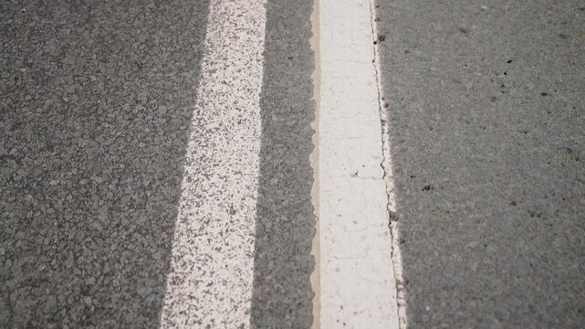 Close-up observation of two parallel white and one solid white road markings on a textured asphalt surface emphasizing transportation infrastructure and urban pavement details