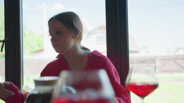 Young brunette woman in crimson shirt sits at wooden table near window, leaning forward searching tabletop for lost item while yard outside glows, ruby drinks and glass pitcher blur in foreground